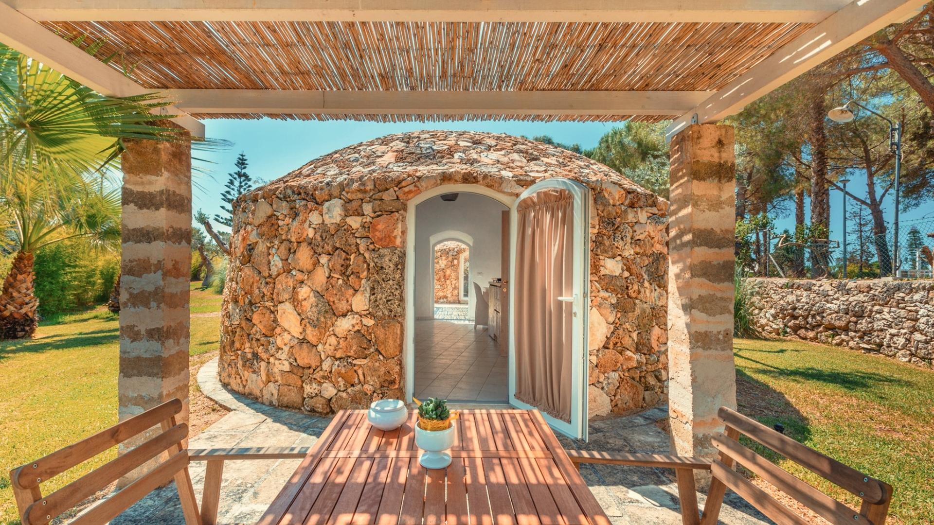 Accogliente trullo in pietra con veranda, tavolo in legno e giardino verde.