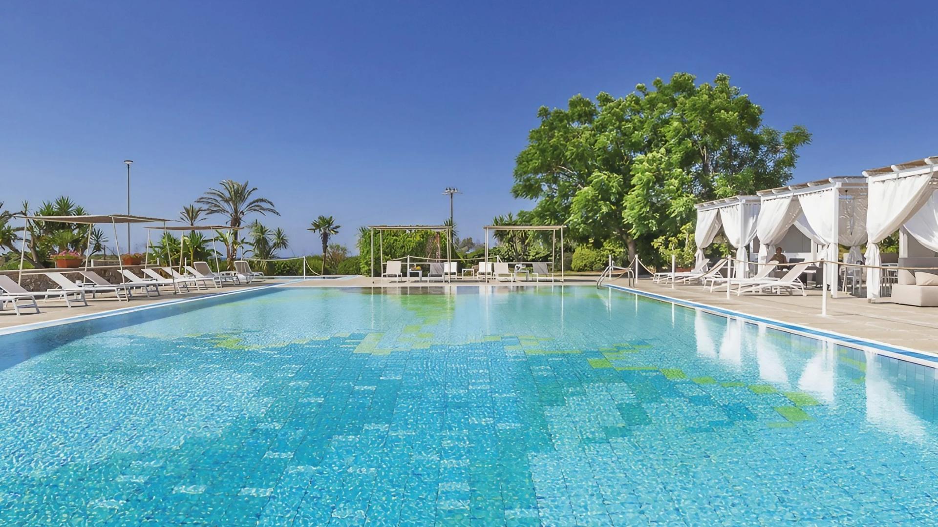 Piscina all'aperto con lettini e gazebo, circondata da palme e alberi verdi.