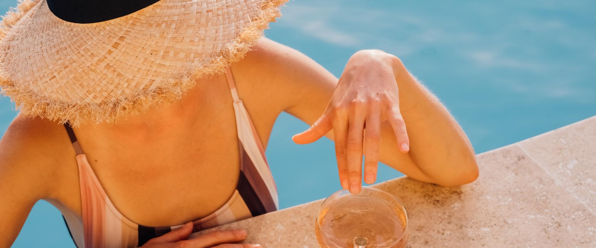 Person with straw hat and swimsuit, by a pool with a drink.