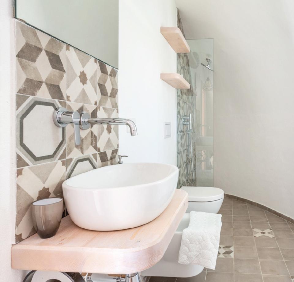 Bagno moderno con lavabo bianco, piastrelle geometriche e mensole in legno.