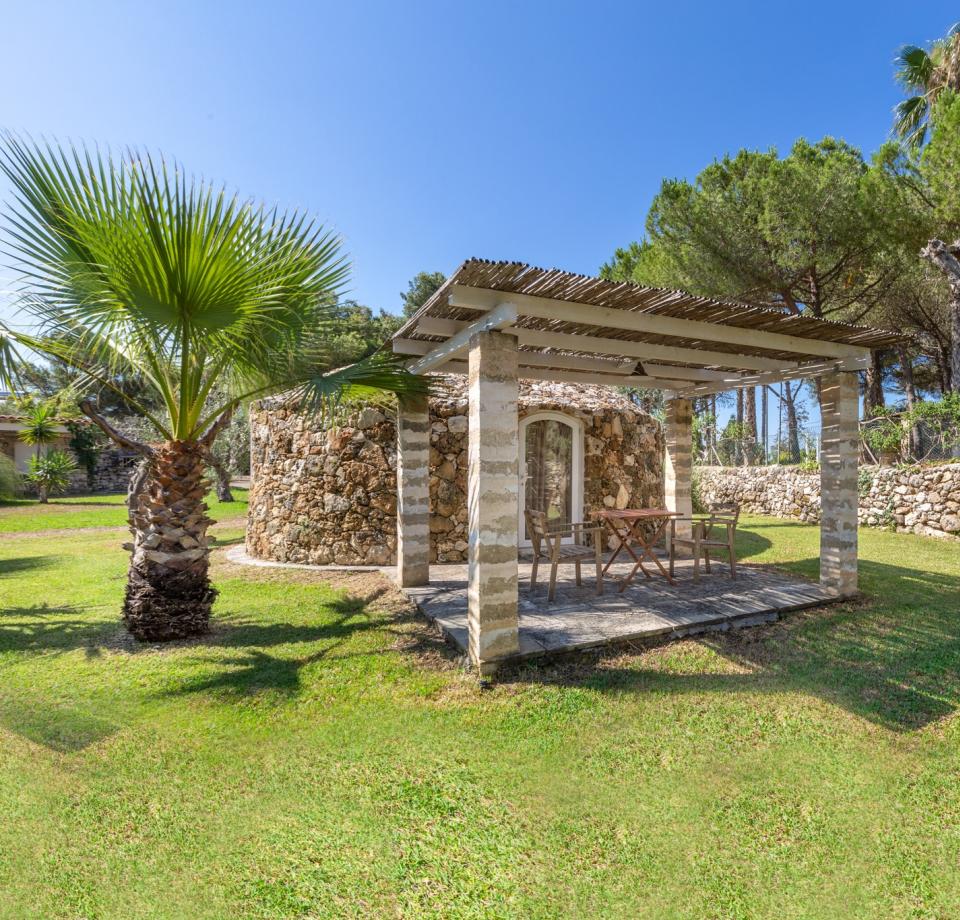 Casetta in pietra con pergolato, circondata da alberi e prato verde.