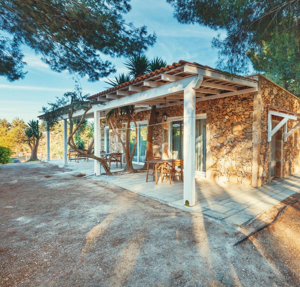 Stone house with white pergola, surrounded by trees and nature.