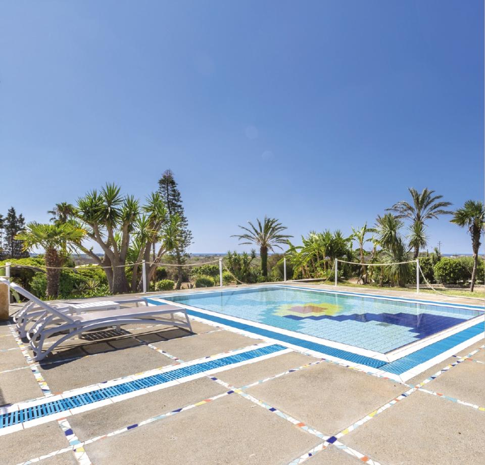 Piscina all'aperto con lettini, circondata da palme e cielo sereno.