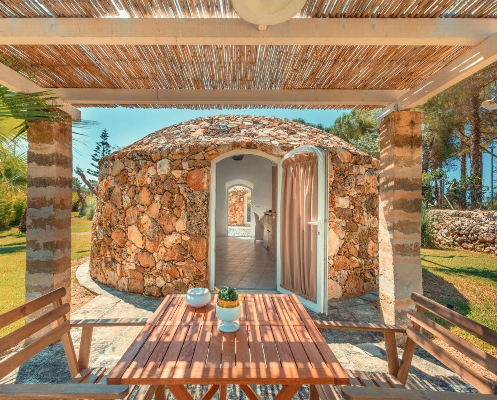 Accogliente trullo in pietra con veranda, tavolo in legno e giardino verde.